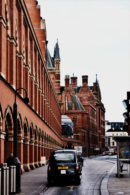 The image shows a narrow urban street adjacent to a large, historic red-brick building with arched windows and decorative brickwork, indicative of Victorian architecture. The building's façade extend