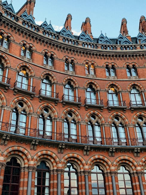 The image shows a narrow urban street adjacent to a large, historic red-brick building with arched windows and decorative brickwork, indicative of Victorian architecture. The building's façade extend